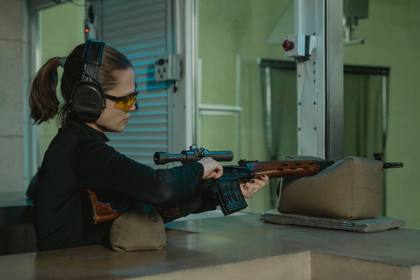 Precision rifle training at an indoor shooting range: woman aiming a scoped rifle while wearing hearing protection, illustrative stock photo