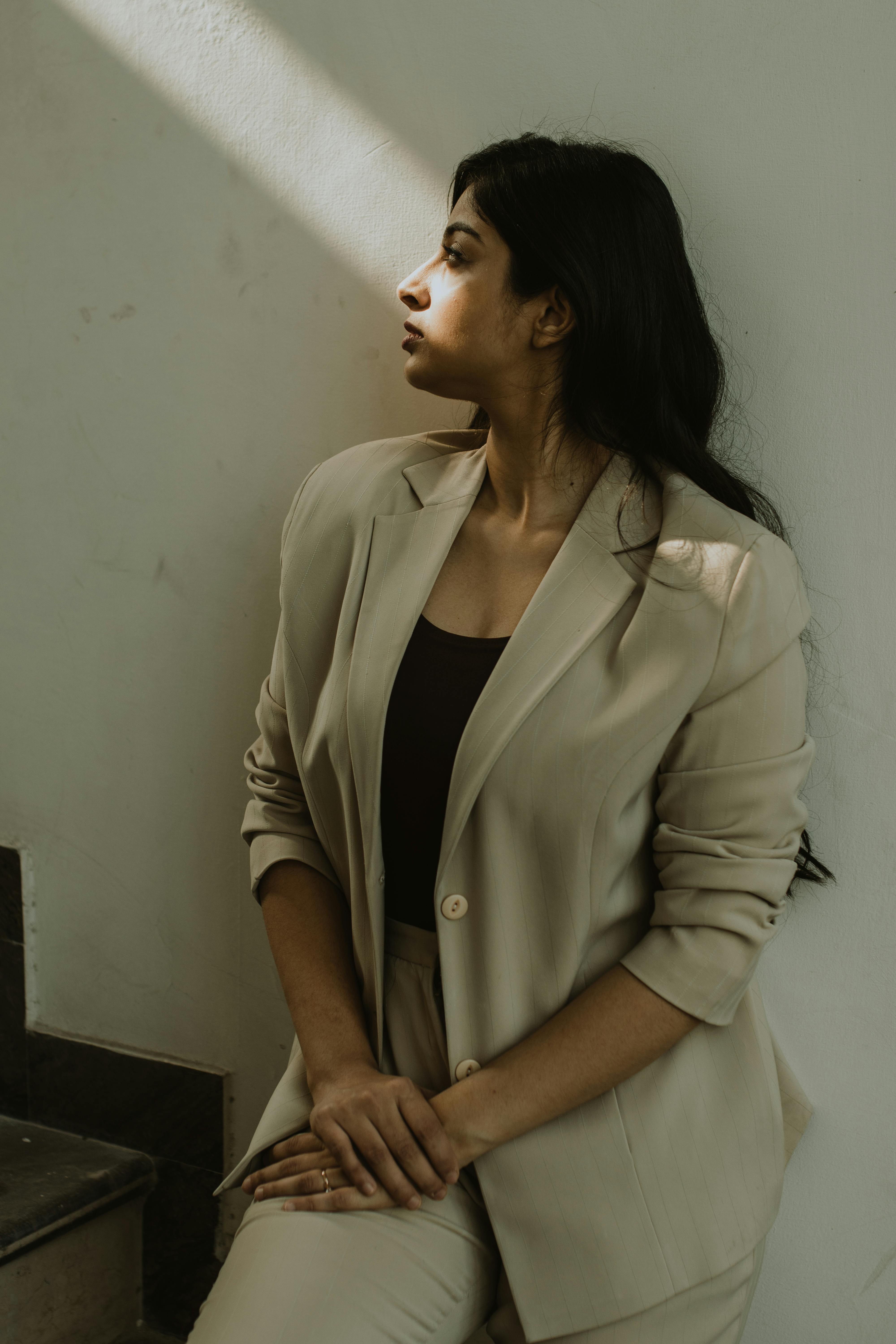 Thoughtful woman in a chic beige blazer sits on a staircase, bathed in sunlight.