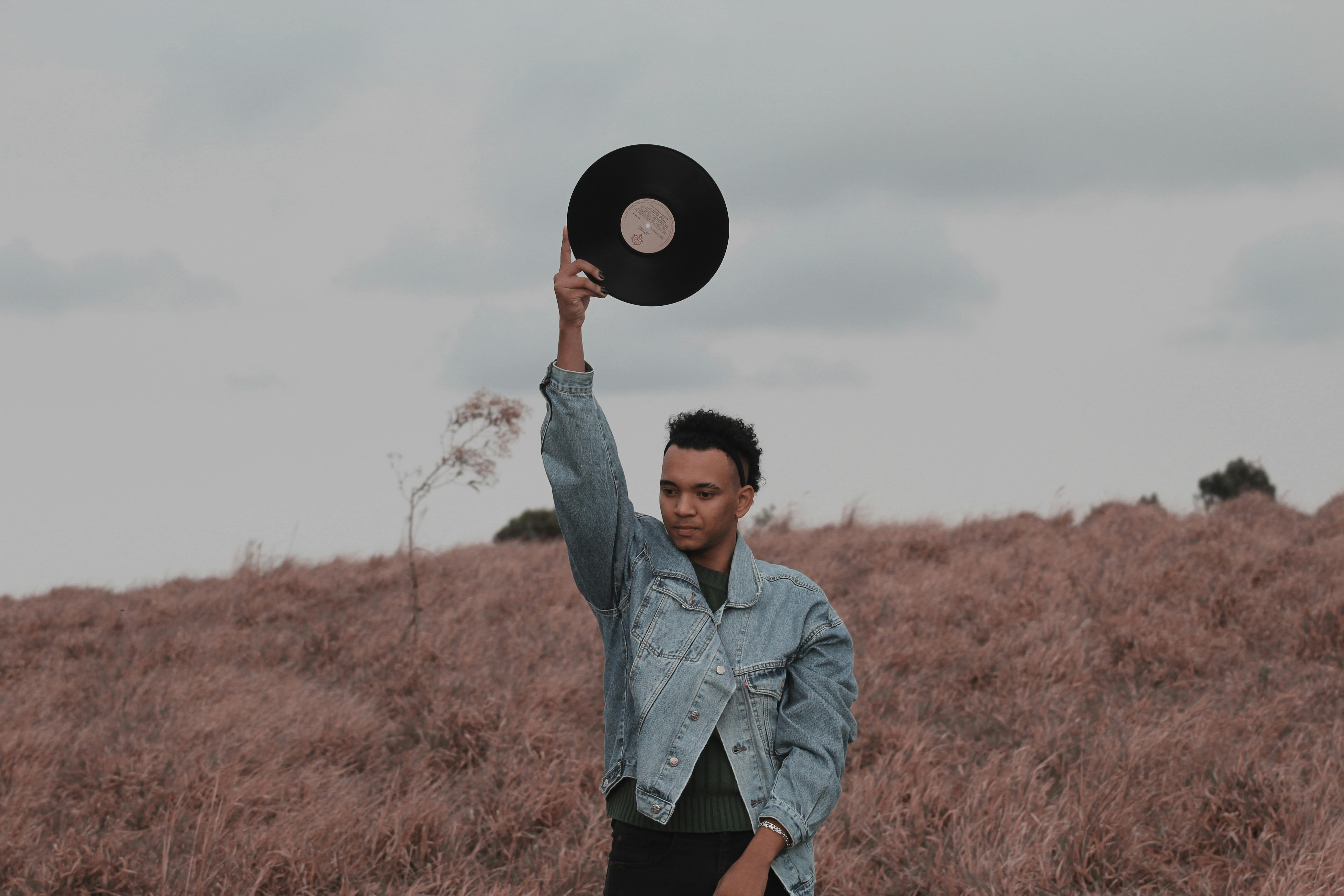 A young man in a denim jacket holds a vinyl record outdoors in a grassy field.