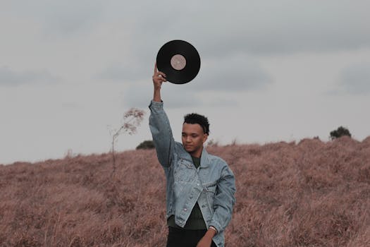 A young man in a denim jacket holds a vinyl record outdoors in a grassy field.