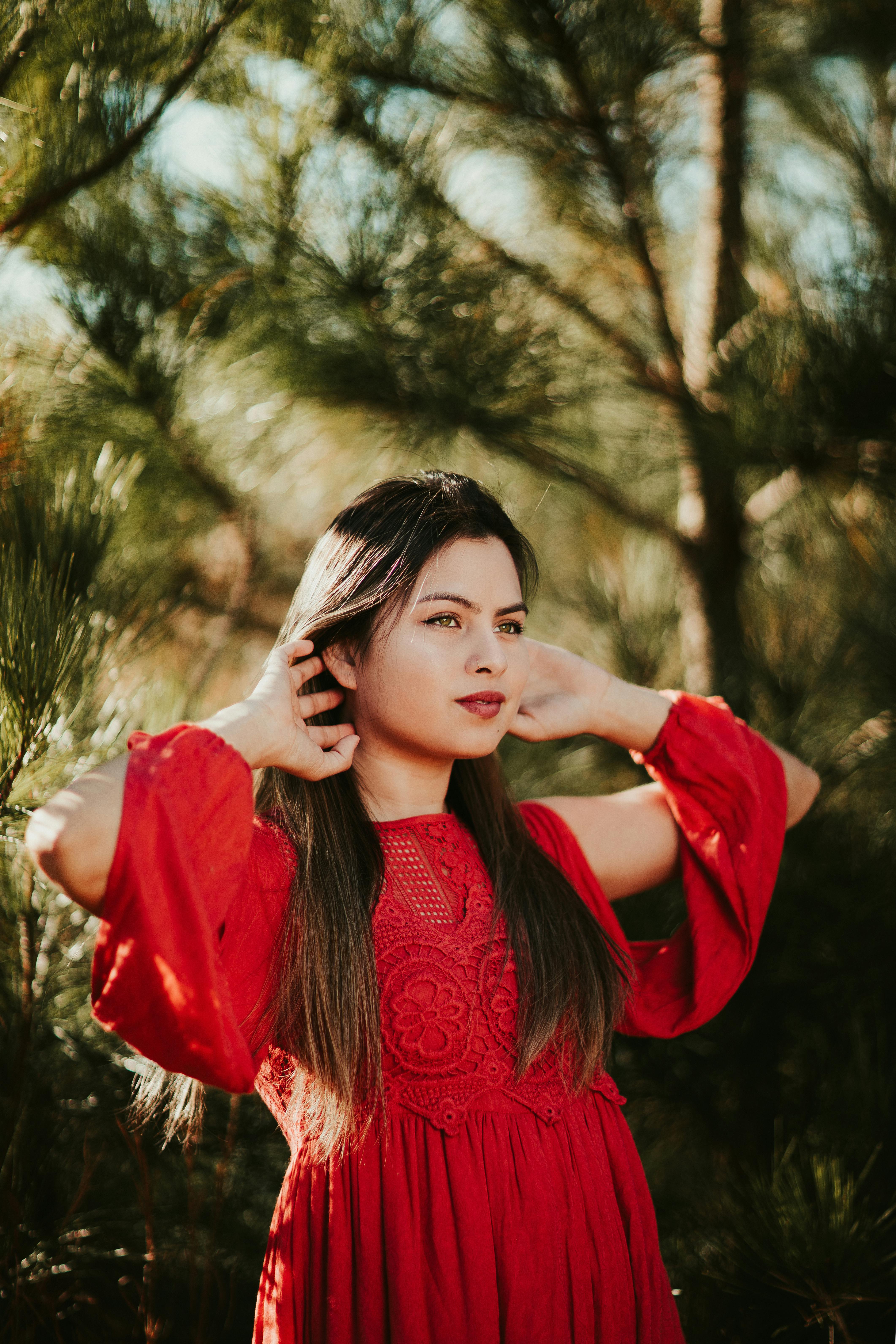 Young female with long hair looking away and touching neck on blurred background of coniferous tree