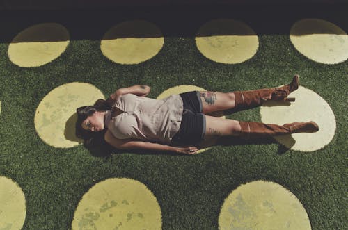 High angle full body of young woman in casual clothes and brown boots lying alone on green grass with yellow circles in sunny summer day and looking away
