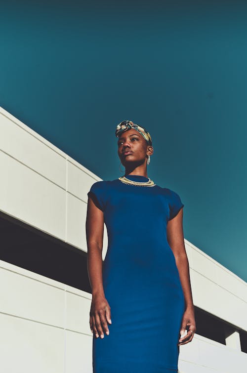 Low angle confident African American woman in blue dress and headband standing near white construction under blue cloudless sky and looking away in sunny day