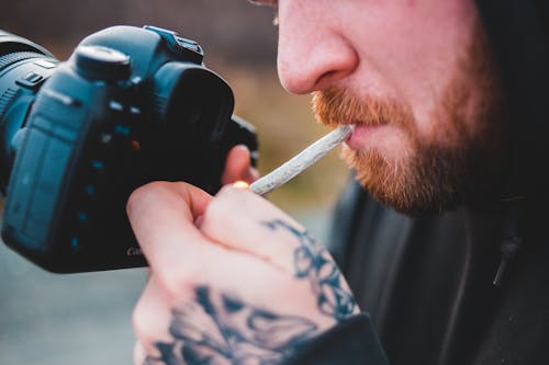Free Side view of crop anonymous tattooed male photographer lighting cigarette in mouth with camera in hand Stock Photo