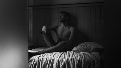Grayscale Photo of a Topless Man Sitting on His Bed