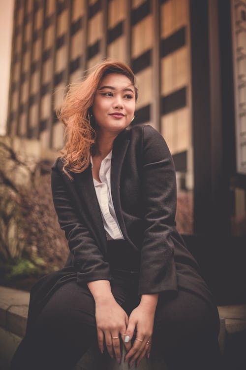 Young stylish Asian lady with dyed hair wearing black jacket smiling while sitting on corner of concrete fence near urban building in daytime