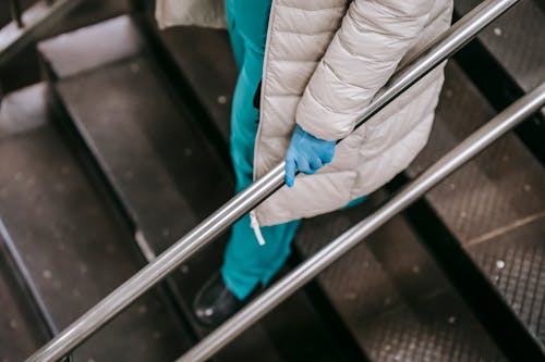 Unrecognizable female wearing medical uniform latex gloves and coat going down on metal stairs leaning on metal handrails