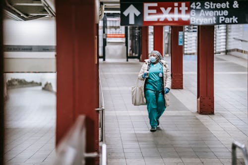 Full body of female in medical uniform protective mask and gloves walking on subway station in New York with paper cup and fabric bag