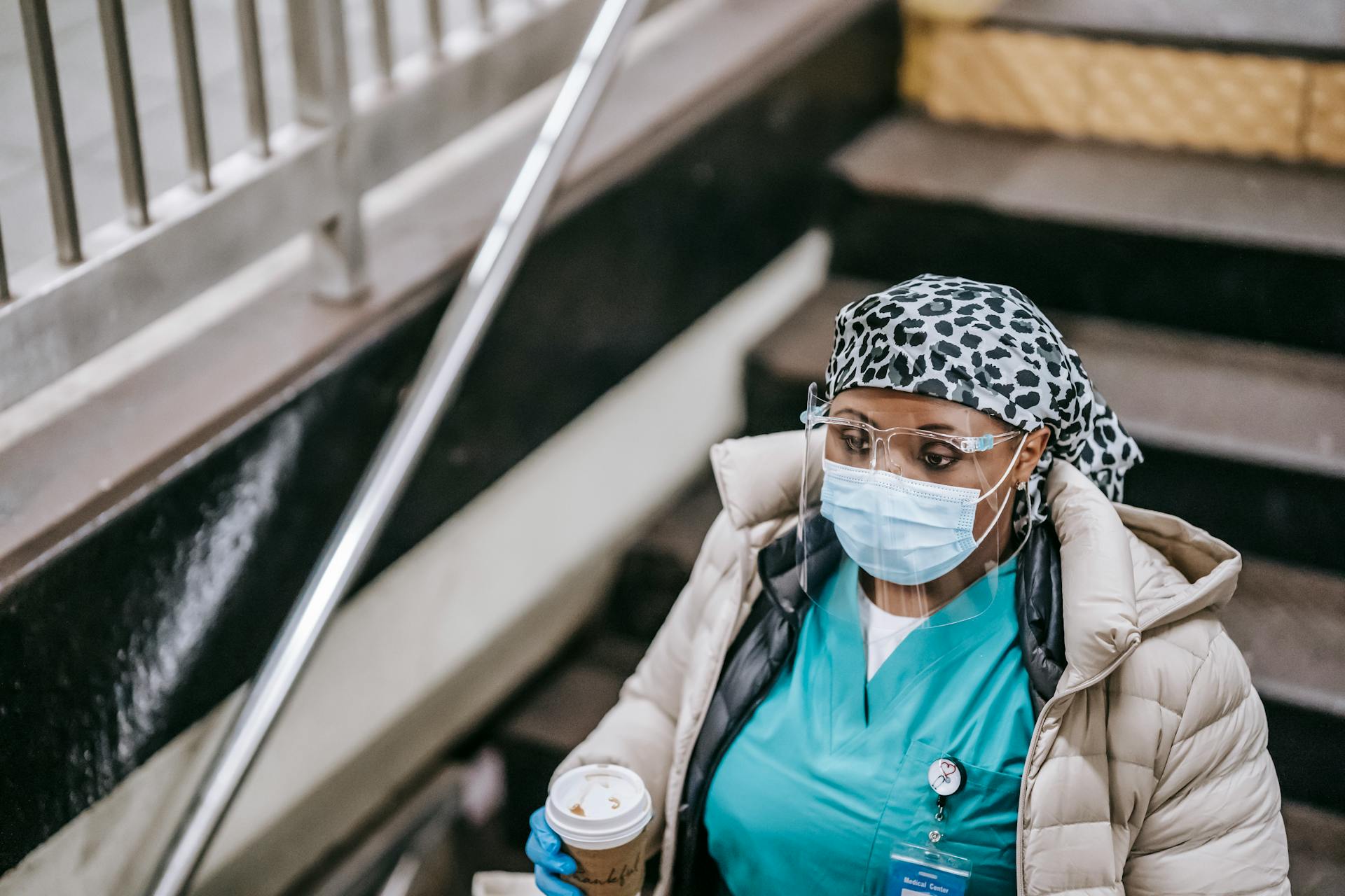 A healthcare worker wearing a mask and gloves descends stairs with a coffee cup.