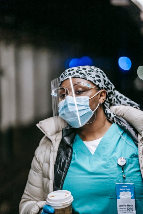 Serious black female nurse with coat and protective masks and with paper cup on blurred background