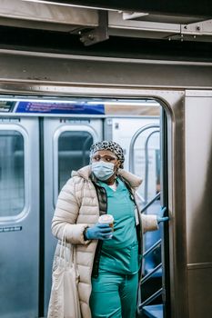 Focused plump African American nurse in uniform and protective mask with coffee to go leaving train in metro station during coronavirus epidemic