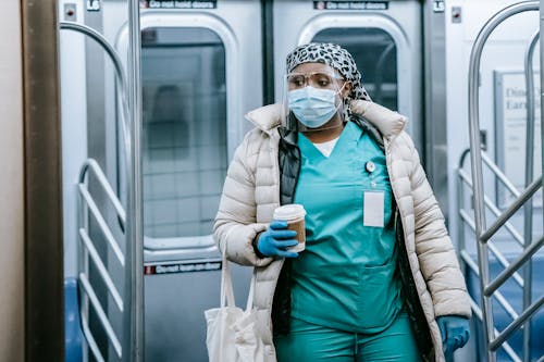 Emotionless African American female doctor in uniform and outerwear wearing protective mask and face shield leaving metro train during coronavirus pandemic