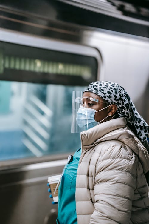 Side view emotionless plump African American female doctor in outerwear wearing mask and shield with takeaway coffee riding metro train and looking away thoughtfully