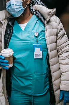 Crop unrecognizable African American nurse in uniform and protective mask wearing warm clothes walking with takeaway coffee in public place