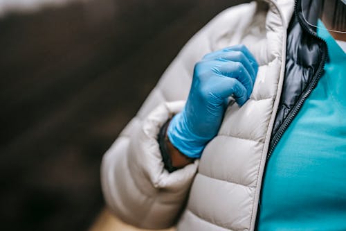 Crop anonymous female doctor in warm clothes wearing uniform and latex gloves standing on blurred background in public place