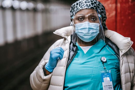 Crop thoughtful African American female doctor in uniform and outerwear wearing protective face mask and face shield standing on metro platform and looking at camera seriously