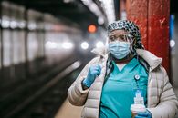 Serious plump African American nurse in uniform and outerwear wearing face mask and latex gloves standing with takeaway coffee standing on metro station platform and looking away thoughtfully