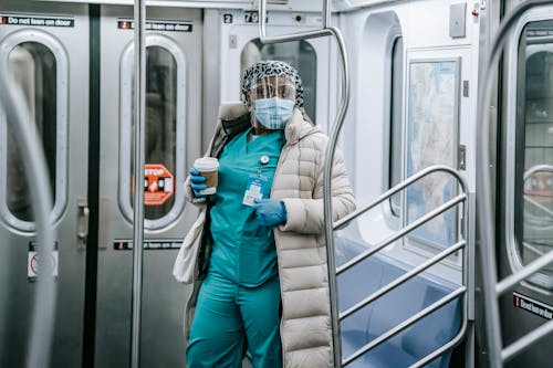 Calm African American female doctor in uniform under outerwear wearing protective mask and face shield standing with takeaway coffee in underground train