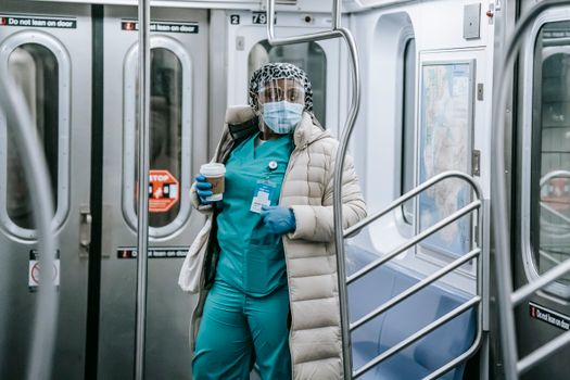 Calm African American female doctor in uniform under outerwear wearing protective mask and face shield standing with takeaway coffee in underground train