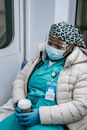Exhausted African American nurse in warm wear and protective face shield mask and gloves sleeping in subway train after long duty during coronavirus pandemic
