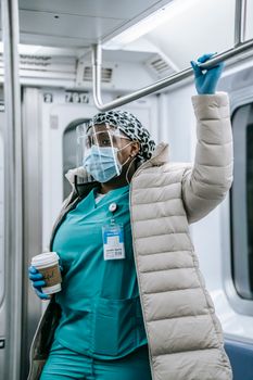Concentrated adult African American nurse in medical uniform under warm clothes wearing mask face shield and gloves holding overhead handle and riding metro train