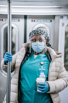 Calm adult African American female doctor in uniform and warm clothes wearing protective face shield mask and gloves grabbing handle and riding metro train during coronavirus epidemic