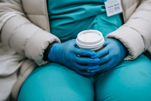 Crop anonymous plump nurse in uniform under warm wear and protective latex gloves sitting in public place and holding takeaway cup