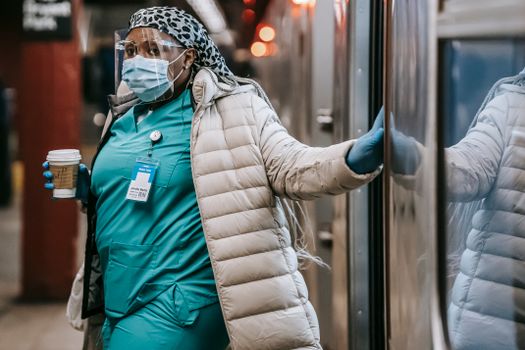 Contemplative African American nurse in uniform and warm clothes wearing medical face shield mask and latex gloves carrying coffee to go and leaving subway train