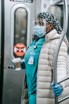 Thoughtful adult African American nurse in medical uniform under warm clothes in protective face shield mask and gloves grabbing handle and riding subway train during coronavirus pandemic
