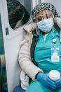 Tired African American nurse in warm clothes and protective mask sleeping in metro train with coffee to go in hands