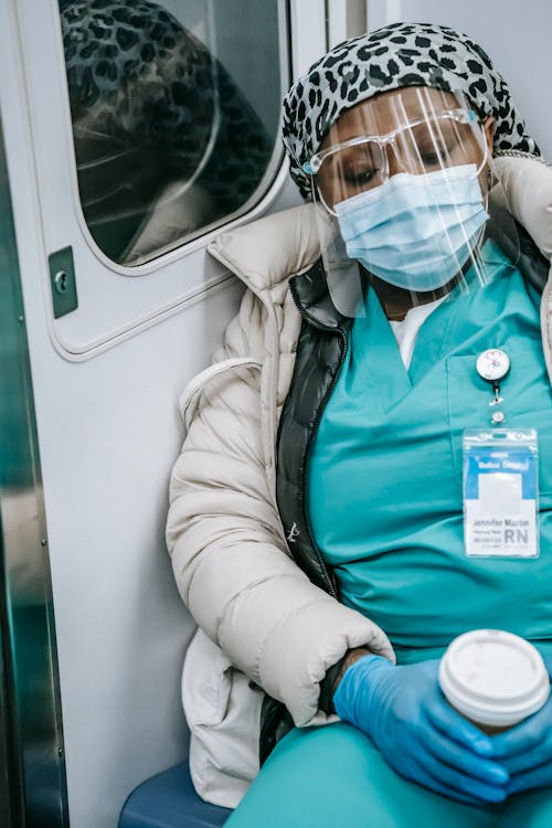 Free Tired African American nurse in warm clothes and protective mask sleeping in metro train with coffee to go in hands Stock Photo