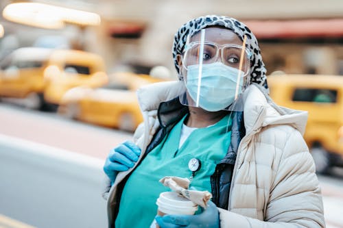 Thoughtful African American nurse in outerwear wearing medical mask face shield and gloves standing on busy street and looking away
