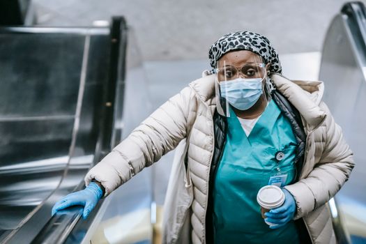 From above contemplative African American nurse in outerwear wearing protective facial shield respirator and gloves riding escalator with takeaway coffee in underground and looking at camera pensively