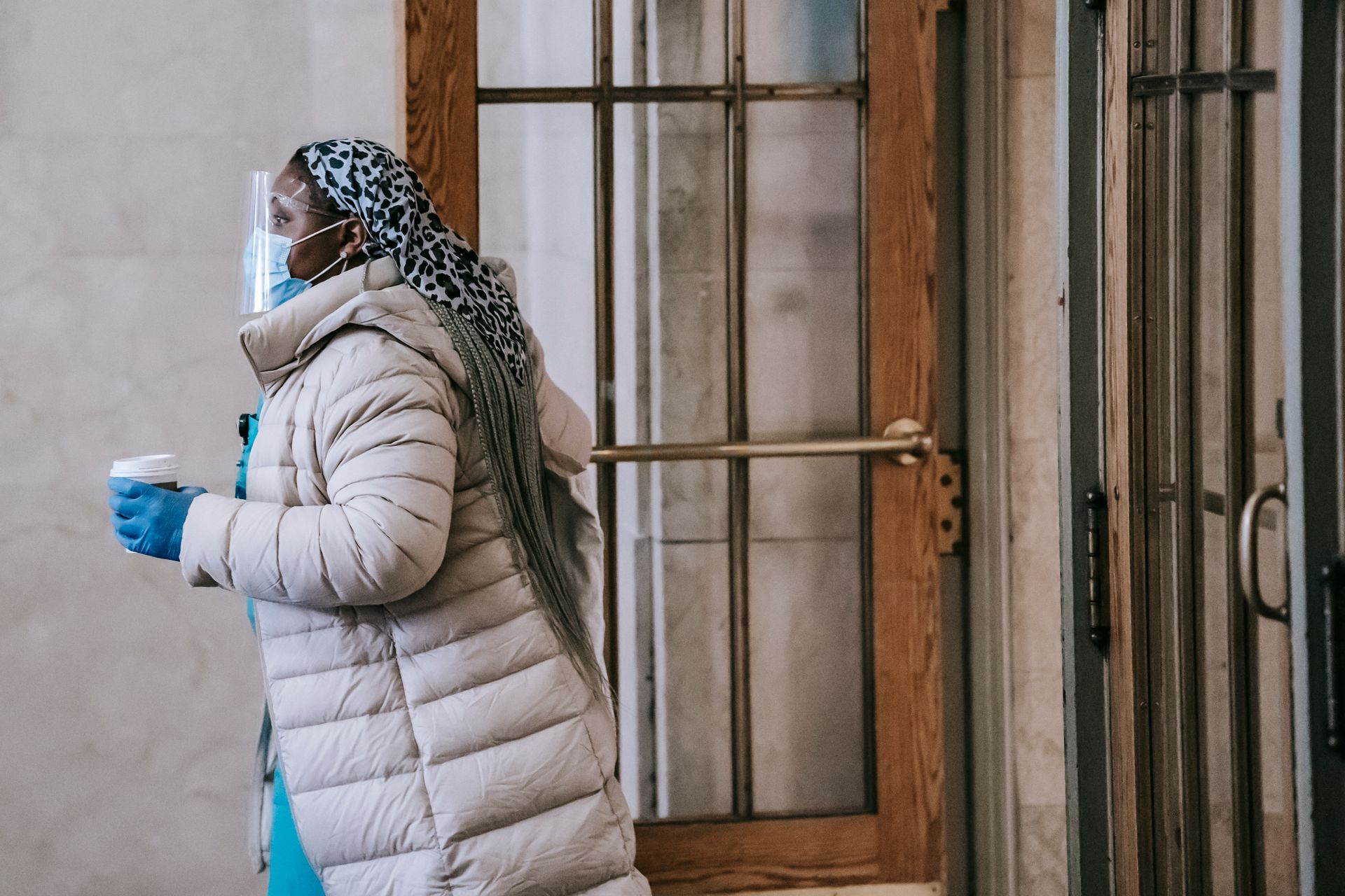 Side view of African American female doctor in face shield with takeaway hot drink looking forward against urban building