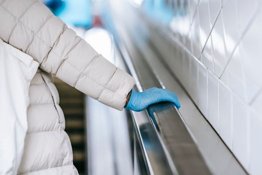 Back view of crop anonymous ethnic female medic in padded coat and latex glove touching handrail of moving stairs in underground