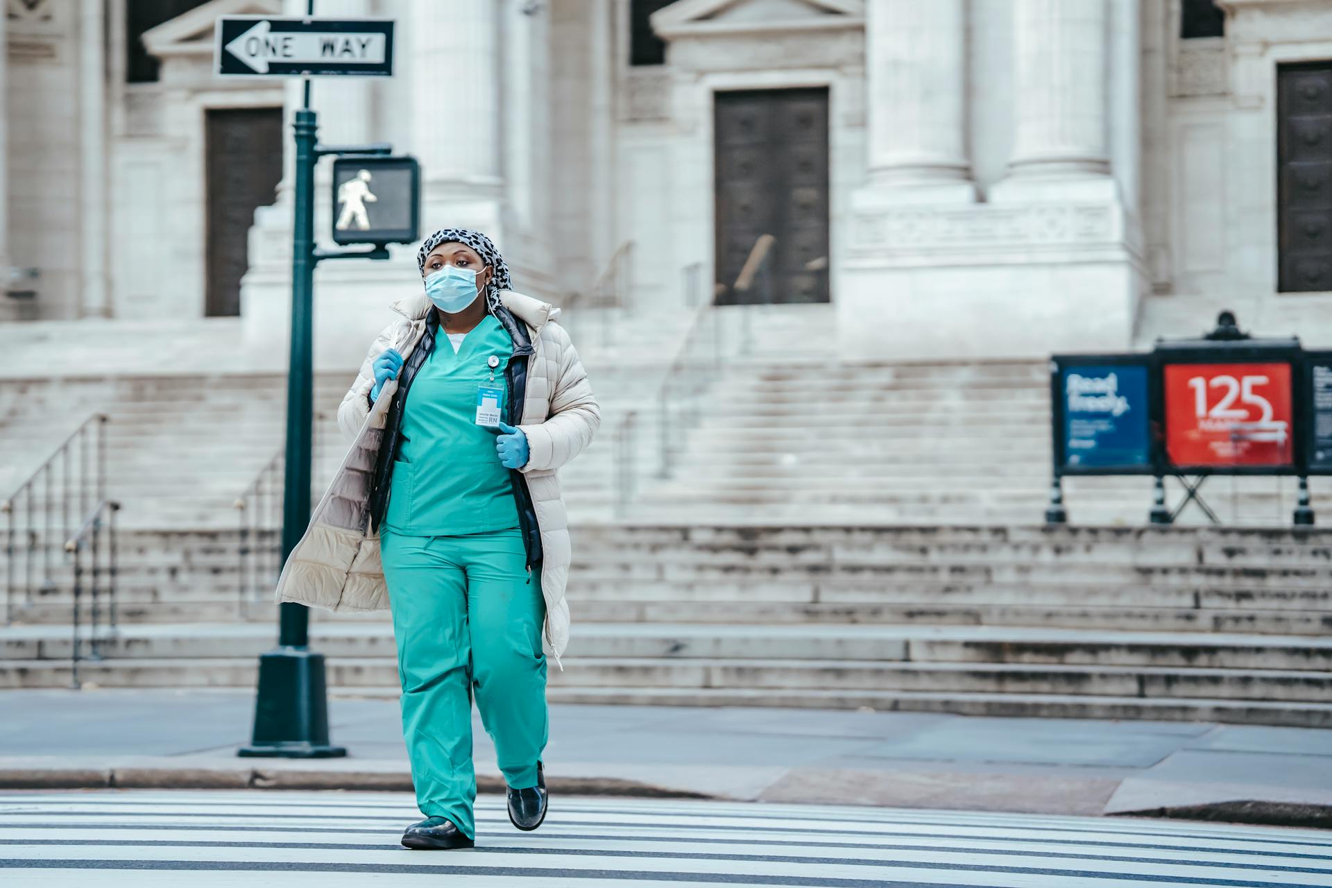 A healthcare worker in scrubs and mask crosses a city street, highlighting urban life during a pandemic.