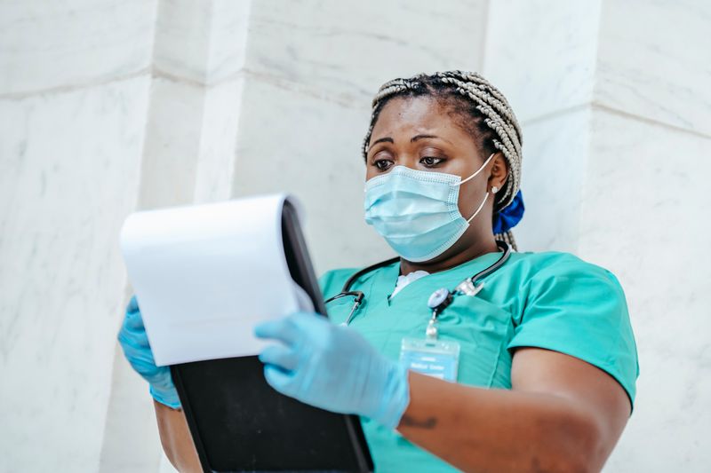 A Black nurse in teal scrubs, a surgical mask and blue gloves, stethoscope around her neck, reads a chart on a clipboard against a marble wall: the health department and clinic staff who deliver WISEWOMAN screenings.