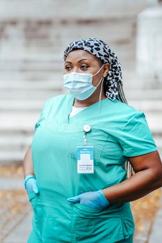 Middle aged African American female medic in sterile mask and gloves with identification card looking away in town