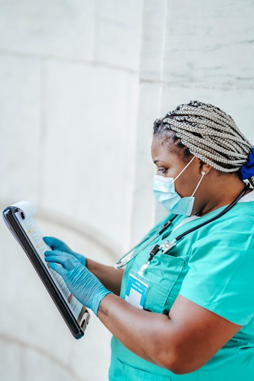 Side view of mature black female medic with Afro braids and clipboard reading text on paper in daytime