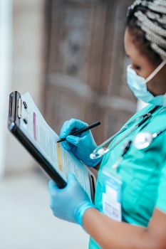 Side view of crop African American female doctor in sterile gloves and mask with clipboard taking notes on paper