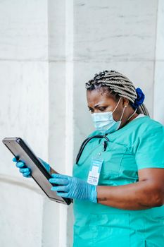 Mature African American female doctor in sterile mask and gloves standing with clipboard in daylight