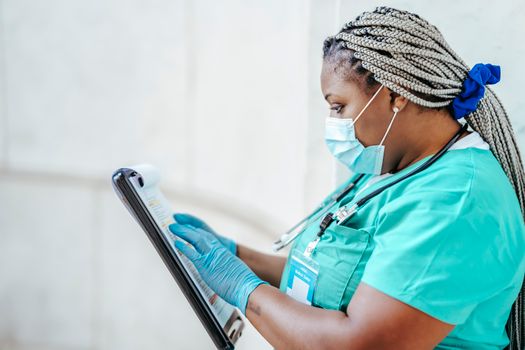 Side view of attentive ethnic female medic with Afro braids in uniform reading paper on clipboard in daytime