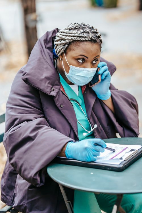 Free Mature ethnic female medic in outerwear and sterile mask sitting at street table with document while talking on cellphone and looking away Stock Photo