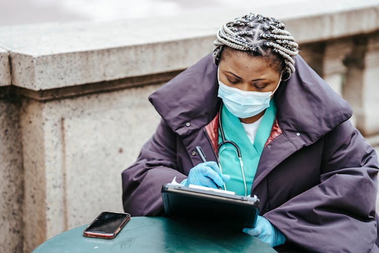 Black Doctor Writing On Paper Near Smartphone In Town