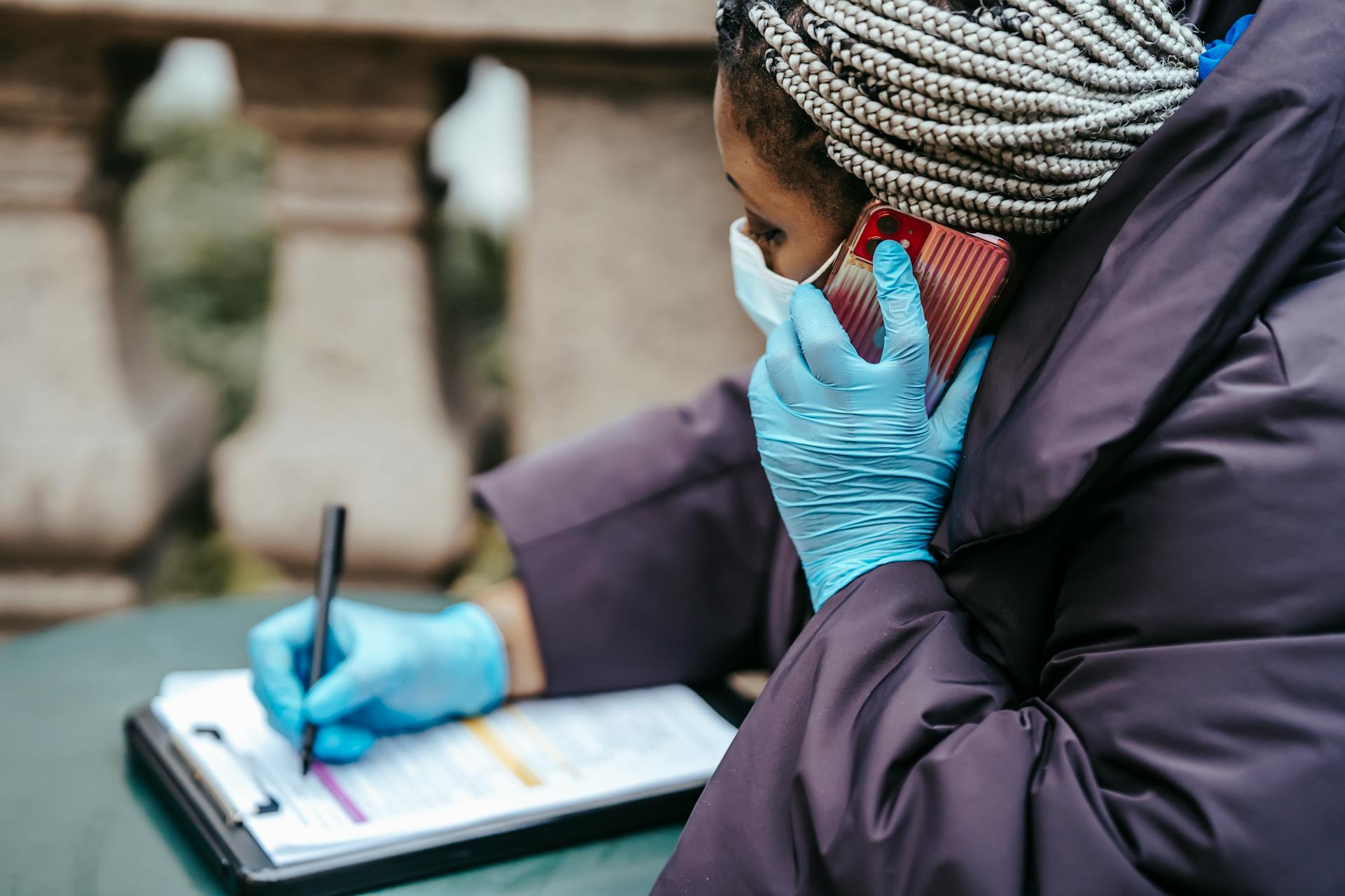 Healthcare professional wearing gloves and a mask writes on a clipboard while speaking on a mobile phone outdoors.