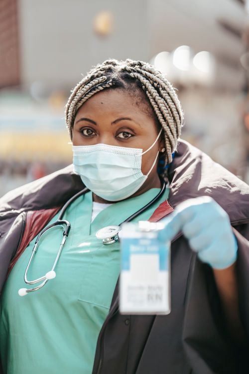 Free Adult black woman nurse in medical clothes and protective mask with gloves and stethoscope standing on street while showing card and looking at camera Stock Photo