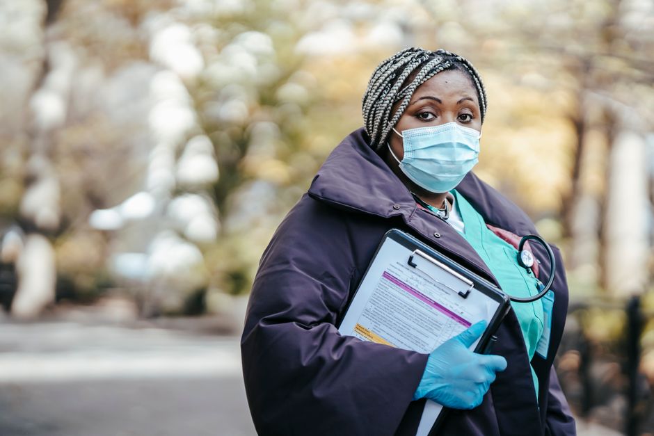 Adult African American woman nurse in medical uniform and protective mask with gloves and stethoscope standing on walkway with papers in clipboard in daytime outside and looking at camera