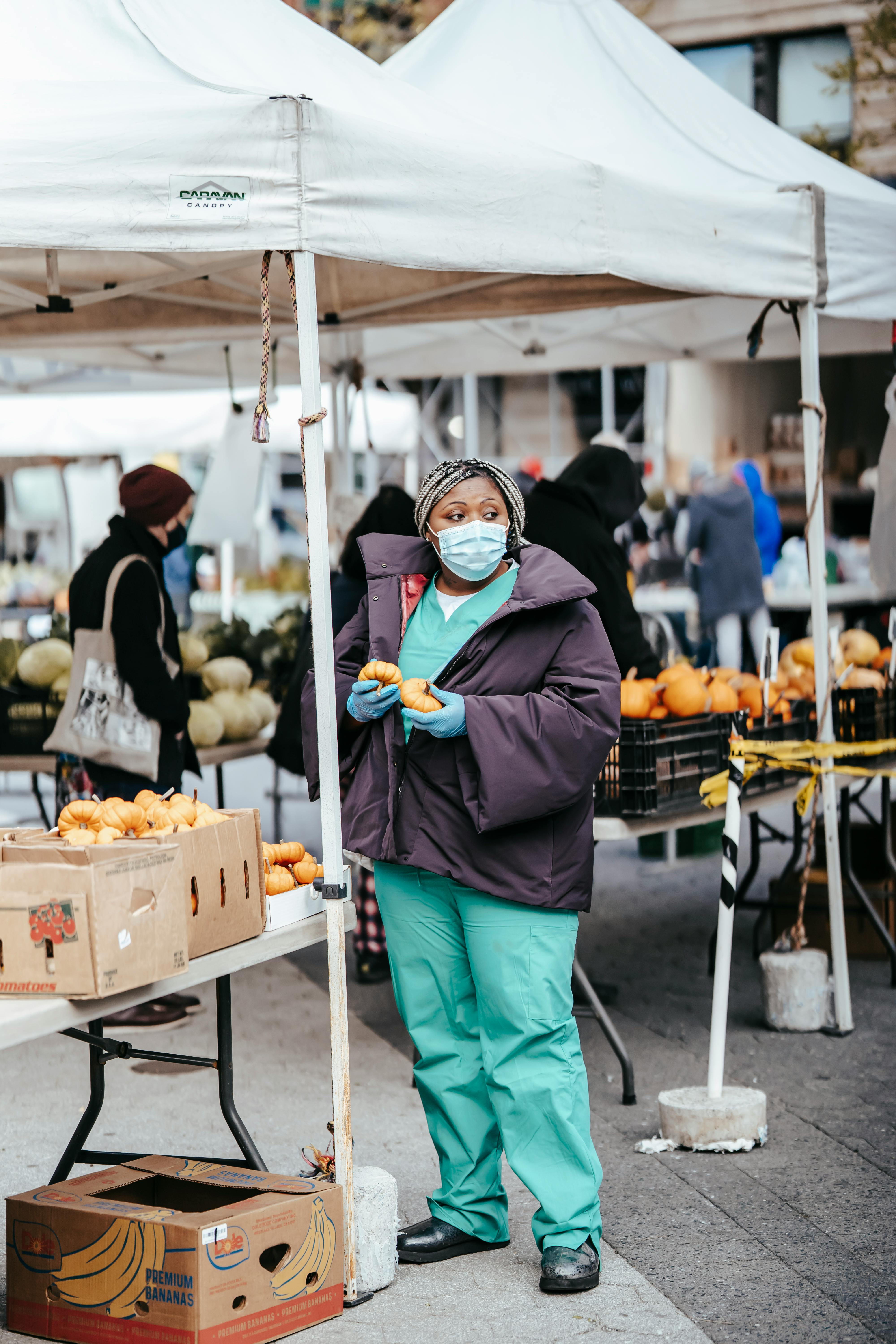 Woman wearing mask and gloves selecting produce at an outdoor market.