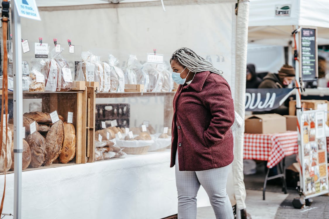 Free Side view of adult African American woman wearing protective mask and warm coat walking in street market while selecting bakery products in daylight Stock Photo