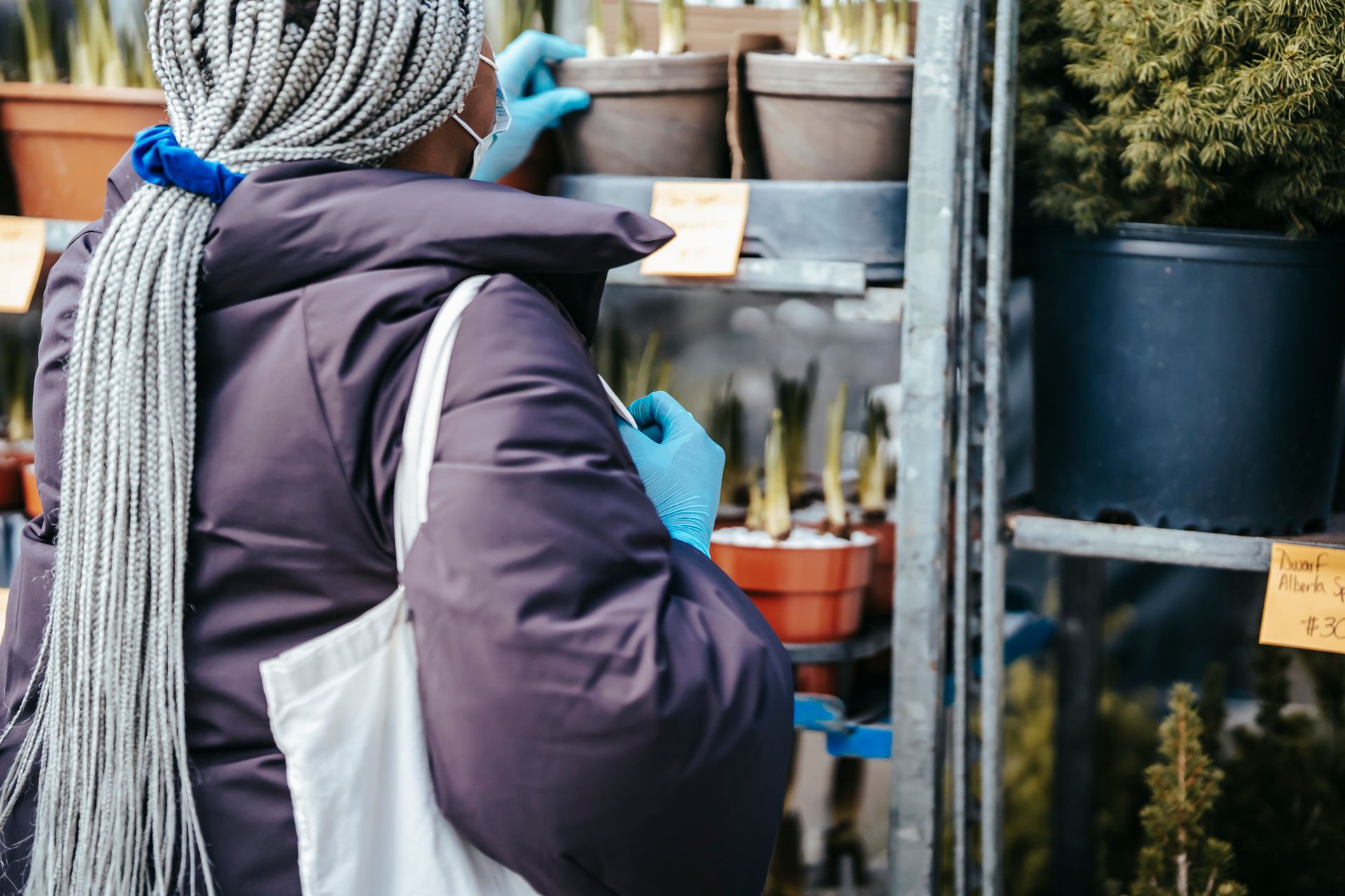A woman wearing gloves and a mask selects potted plants at an outdoor market.